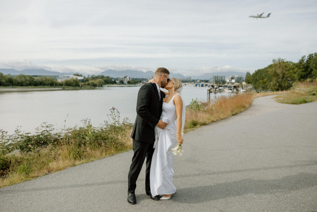Bride and groom kissing outside of the UBC Boathouse Lower Mainland Wedding Venue with a plane flying overehead