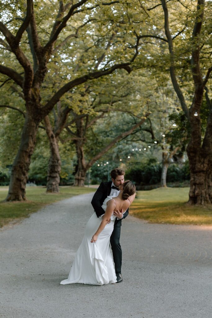 Groom is dipping bride and going for a kiss along a tree lined path outside of the Stanley Park Pavilion 